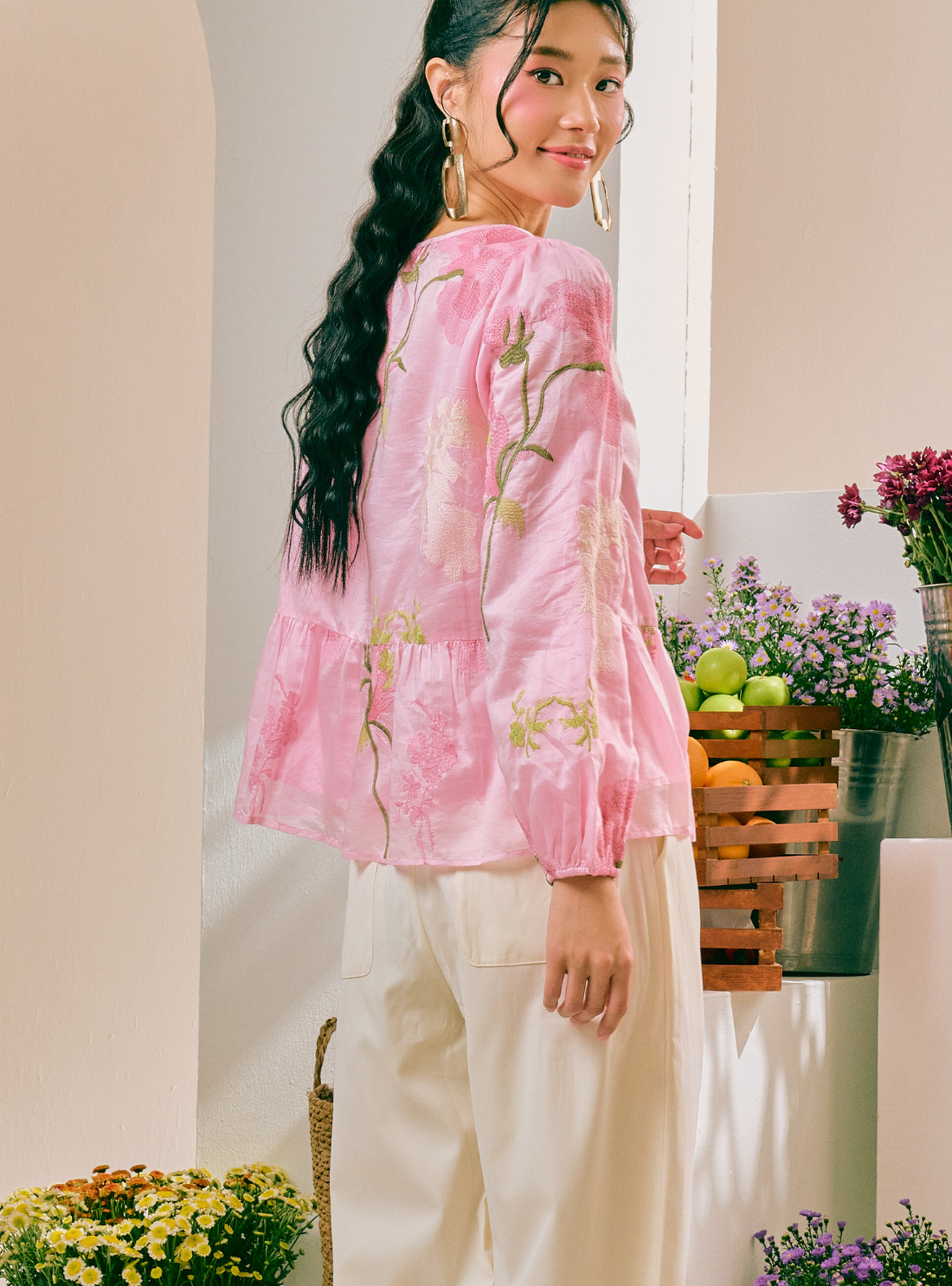 A woman wearing a light pink embroidered blouse with green and yellow floral details, paired with cream-colored pants, standing in a brightly lit indoor setting with potted plants and fruit baskets.