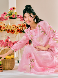 A woman wearing a pink long-sleeve dress with floral embroidery sits in a studio setting decorated with potted flowers and a wicker basket.