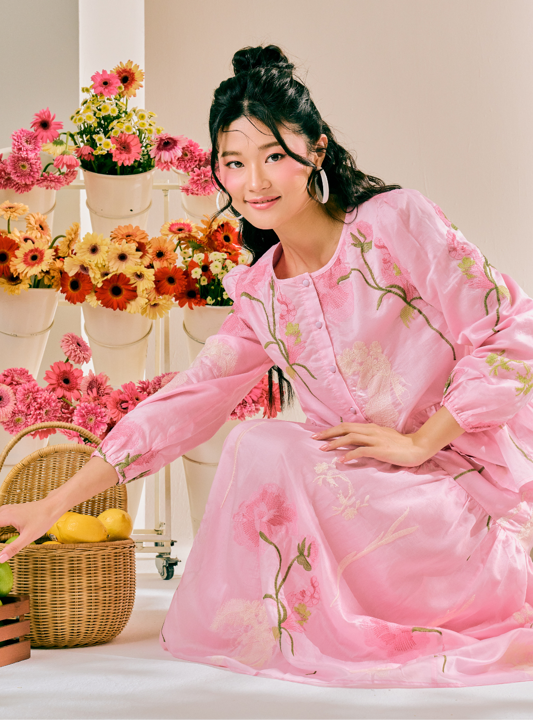 A woman wearing a pink long-sleeve dress with floral embroidery sits in a studio setting decorated with potted flowers and a wicker basket.
