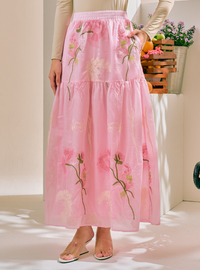 A model wearing a tiered pink maxi skirt with embroidered pink flowers and green stems, paired with a beige long-sleeve top and clear high-heel sandals.