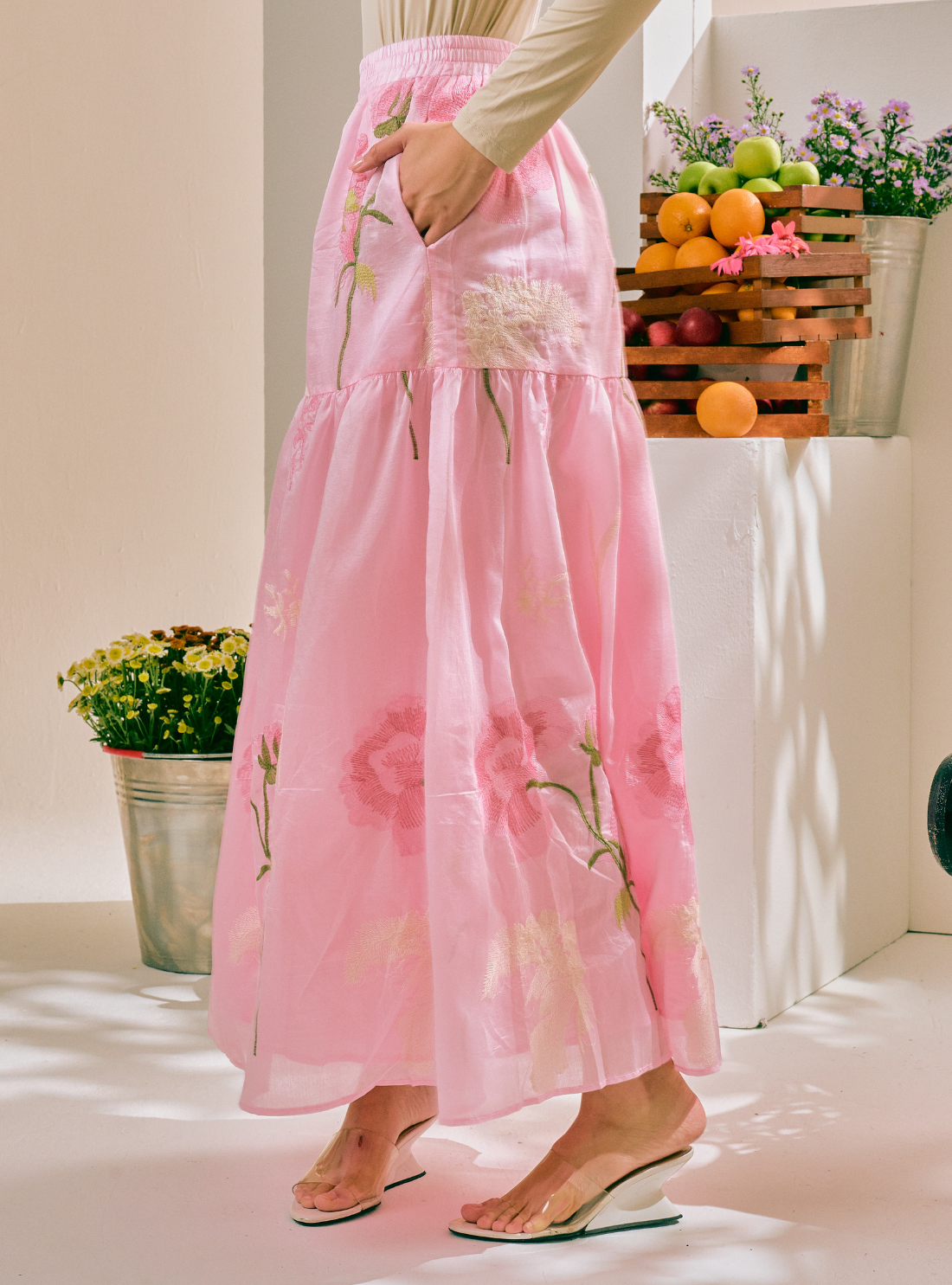 A woman wearing a long, tiered pink maxi skirt with floral embroidery and a matching pink top, standing in a bright indoor setting with plants and fruit crates in the background.