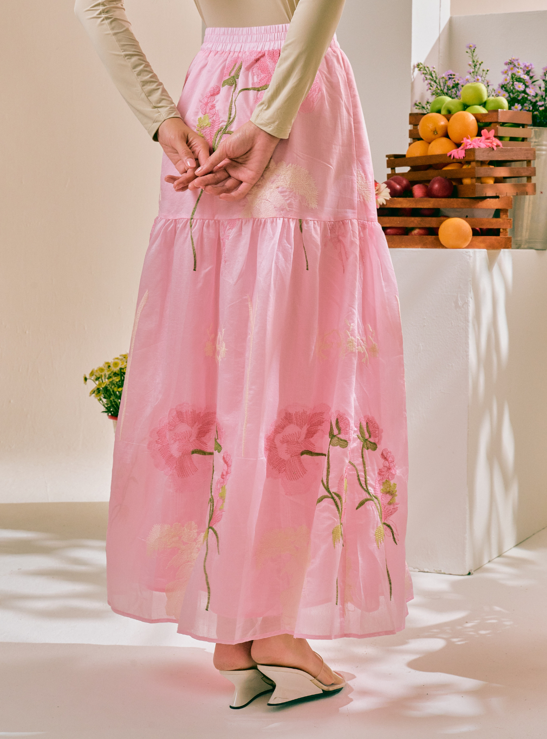 A model wearing a long, tiered pink skirt with large floral embroidery, paired with a cream long-sleeve top and white heels, standing in a brightly lit studio with decorative fruit crates in the background.