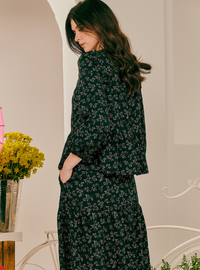 A woman wearing a dark green long-sleeve button-up shirt and matching long skirt with a small floral print, photographed in an indoor setting with a vase of yellow flowers on the left.