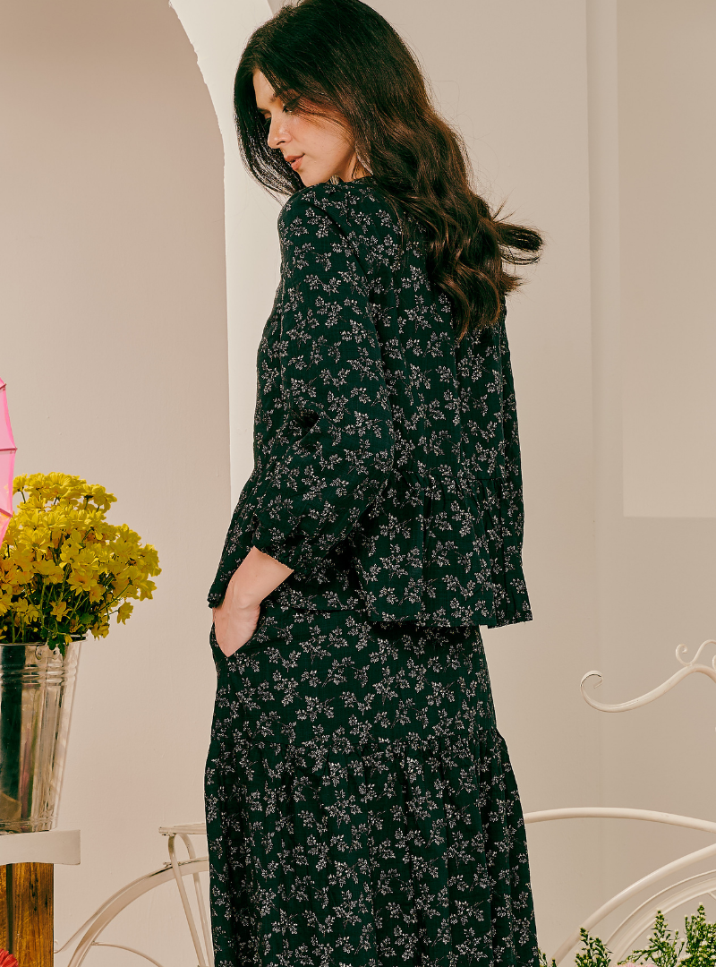 A woman wearing a dark green long-sleeve button-up shirt and matching long skirt with a small floral print, photographed in an indoor setting with a vase of yellow flowers on the left.