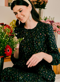 A woman wearing a black long-sleeve floral pajama set with a small multicolor print, sitting in a cozy indoor setting surrounded by colorful flower arrangements.