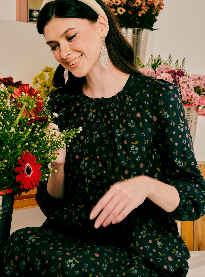 A woman wearing a black long-sleeve floral pajama set with a small multicolor print, sitting in a cozy indoor setting surrounded by colorful flower arrangements.