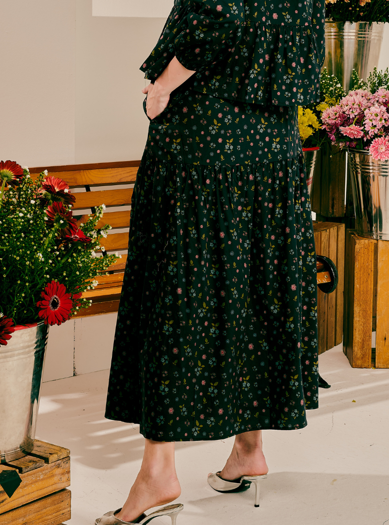 A woman wearing a dark green midi dress with a small pink and white floral print, standing in a styled indoor setting with flower arrangements and wooden crates.
