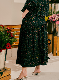 A woman wearing a dark green midi dress with a small pink and white floral print, standing in a styled indoor setting with flower arrangements and wooden crates.