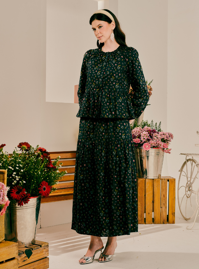 A woman models a dark green two-piece floral set consisting of a long-sleeve peplum top and a matching maxi skirt, styled with a black headband and metallic heels in a bright studio setting with flowers and a wooden bench.