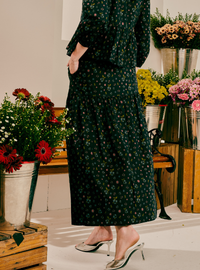A woman wearing a dark green floral print maxi dress with three-quarter sleeves and silver pointed-toe mule heels, standing in a flower shop surrounded by potted flowers.